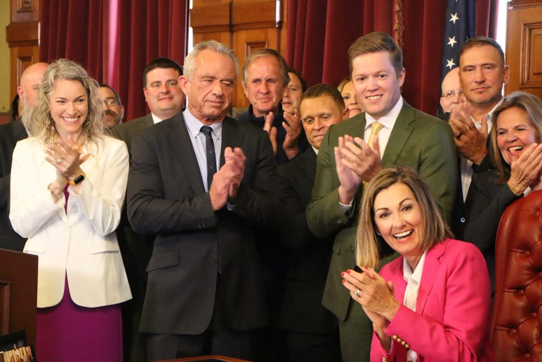 Austin Harris, in the green suit, applauding beside HHS Secretary Robert F. Kennedy Jr. as Gov. Kim Reynolds signs a bill, Iowa Capitol